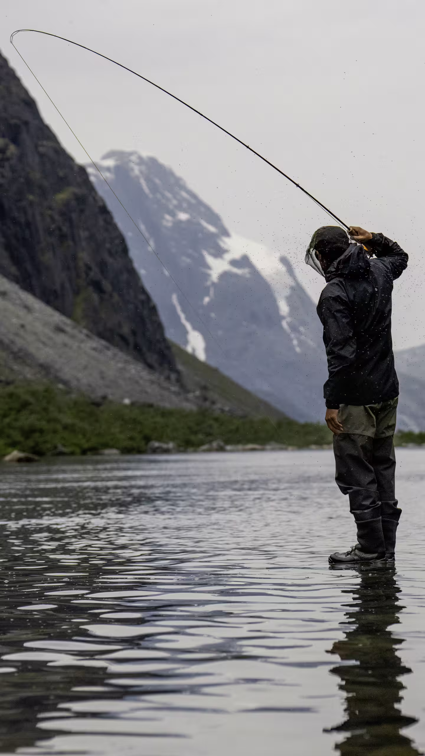 Person fly fishing in a river with mountain landscape and snow in the background.