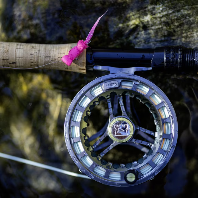 Close-up of a fishing reel attached to a rod with a bright pink fishing fly tied near the wooden handle.
