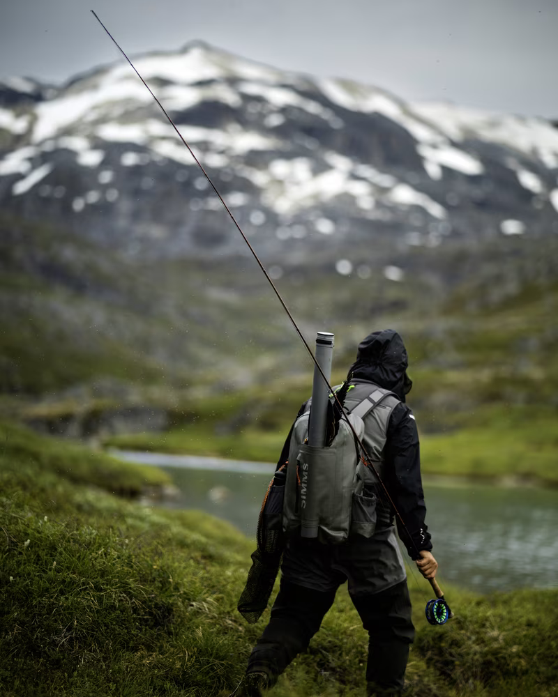 Person fishing near a river in a mountainous landscape with snow patches on peaks.