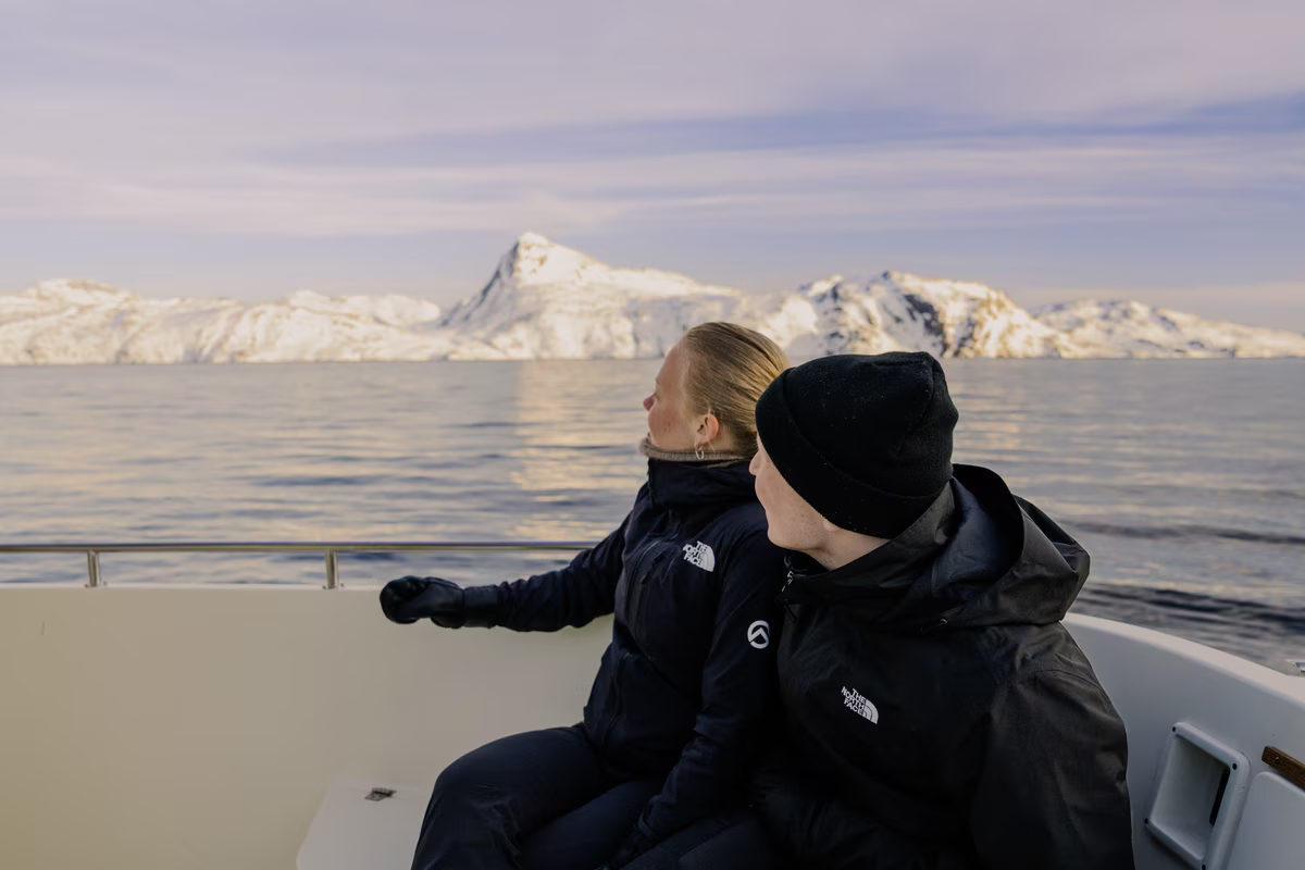 Two people in black jackets on a boat looking at snowy mountains across calm water.