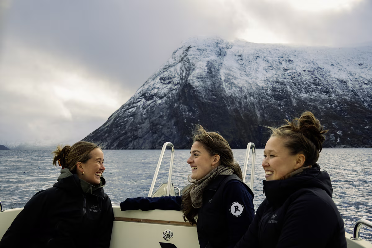 Three women smiling and chatting on a boat with snowy mountain and cloudy sky in the background.