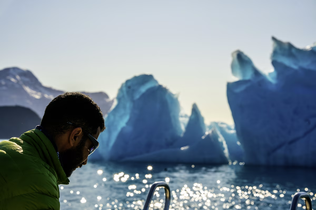 Man with sunglasses on boat looking at large blue icebergs in sparkling cold water.