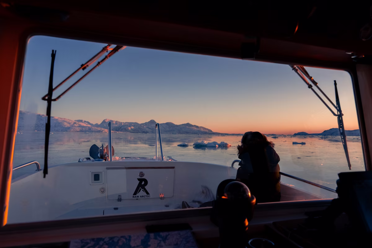 View from boat cabin of person sitting on deck with icy waters and mountains at sunset.