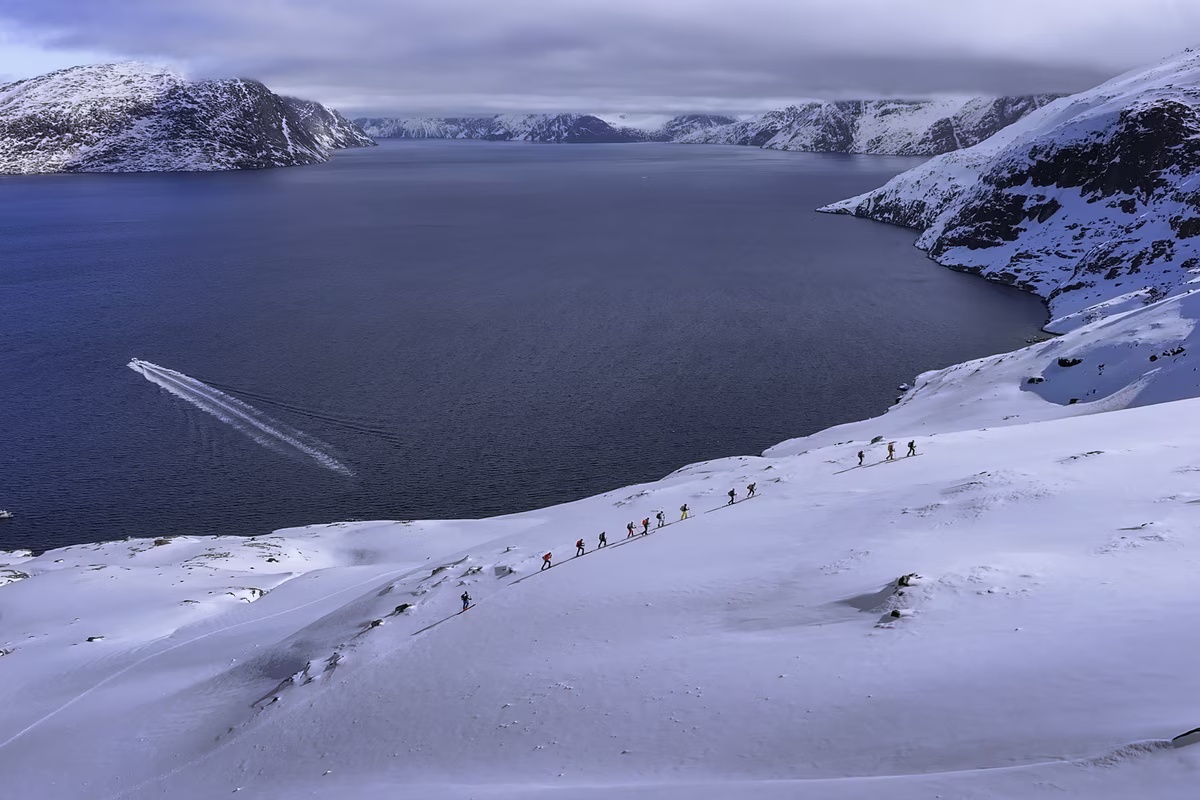 Group of hikers trekking in a snowy landscape beside a large dark blue fjord under cloudy sky.