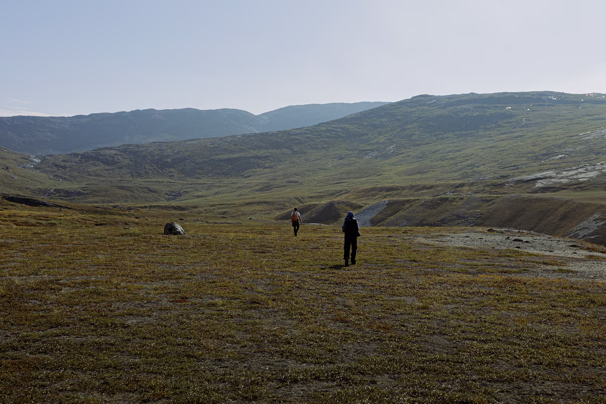 Two hikers walking on a vast mossy plain with rolling green hills under a clear sky.
