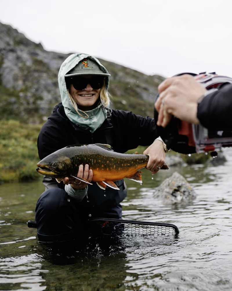Person in waders holding a colorful fish in a shallow stream as another takes a photo.
