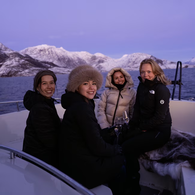 Four women in winter coats sitting on a boat with snow-covered mountains and a lake in the background.