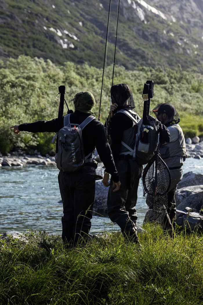 Three people with fishing gear standing by a river, surrounded by greenery and mountains.