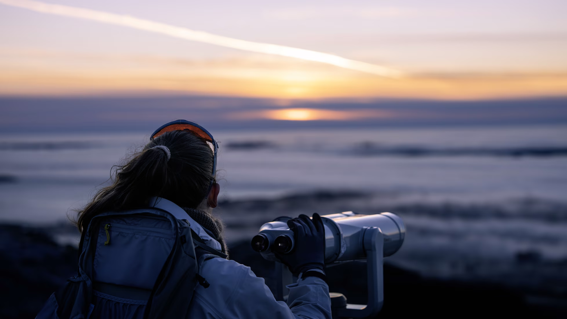 Person with a ponytail using binoculars at a rocky beach during sunset.
