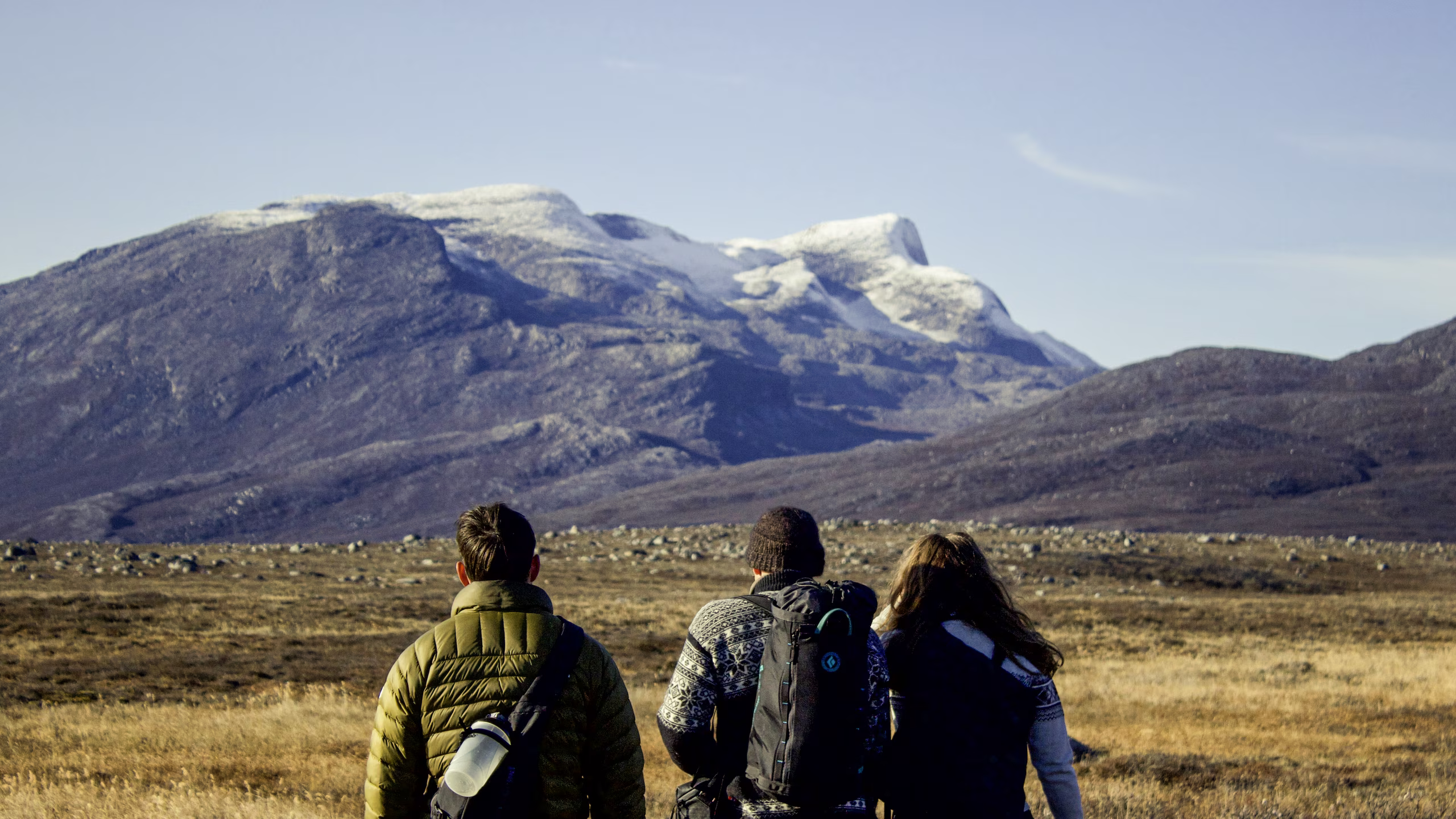 Three hikers walking toward snow-capped mountains in a grassy, rocky landscape under a clear sky.