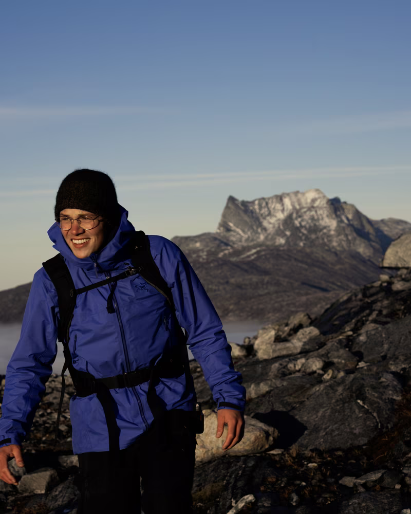 Smiling hiker in a blue jacket and black hat walks on rocky terrain with snowy mountains behind.