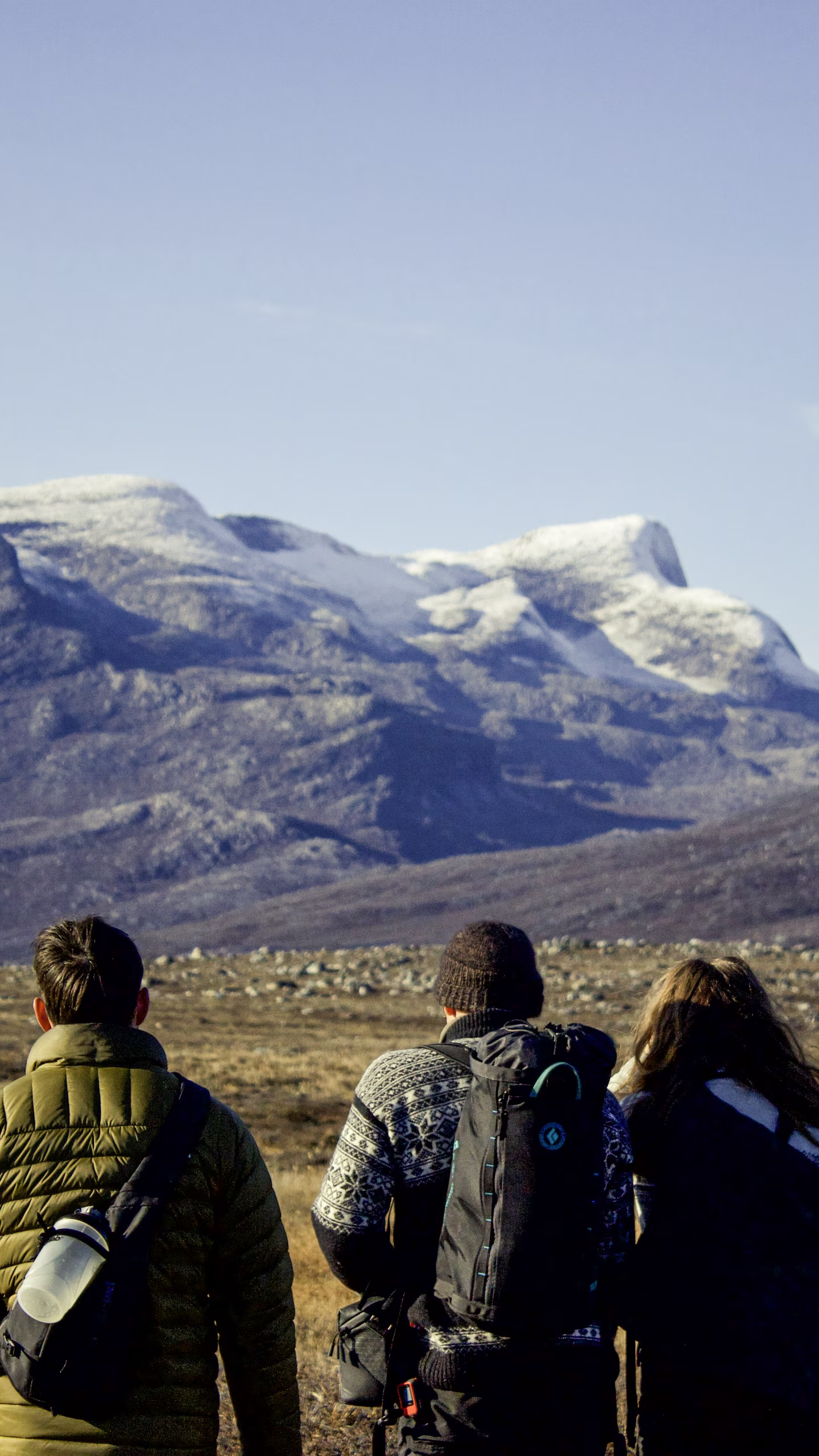 Three hikers with backpacks looking at distant snow-capped mountains under a clear sky.