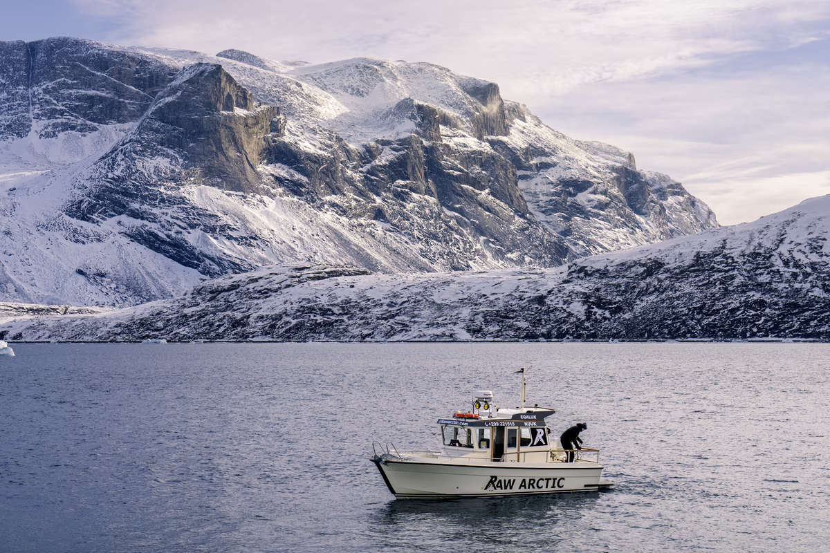 Boat named Raw Arctic with a person onboard in cold water near snowy rocky mountains.