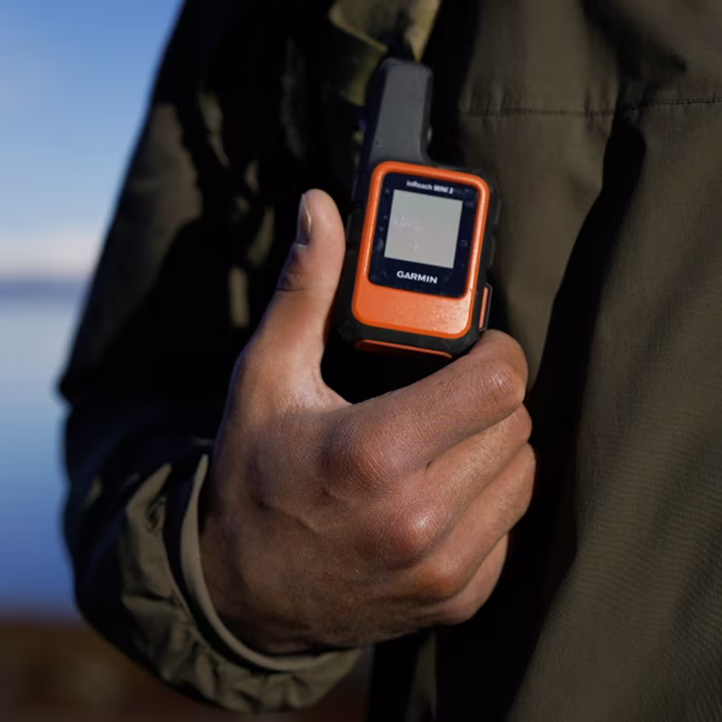 Hand holding an orange Garmin inReach Mini 2 satellite communicator device, person wearing dark jacket.