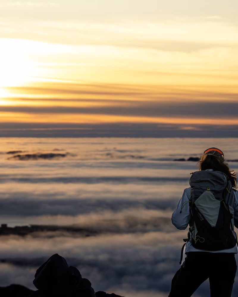 Hiker with backpack watches sunset above a sea of clouds from a mountain peak.
