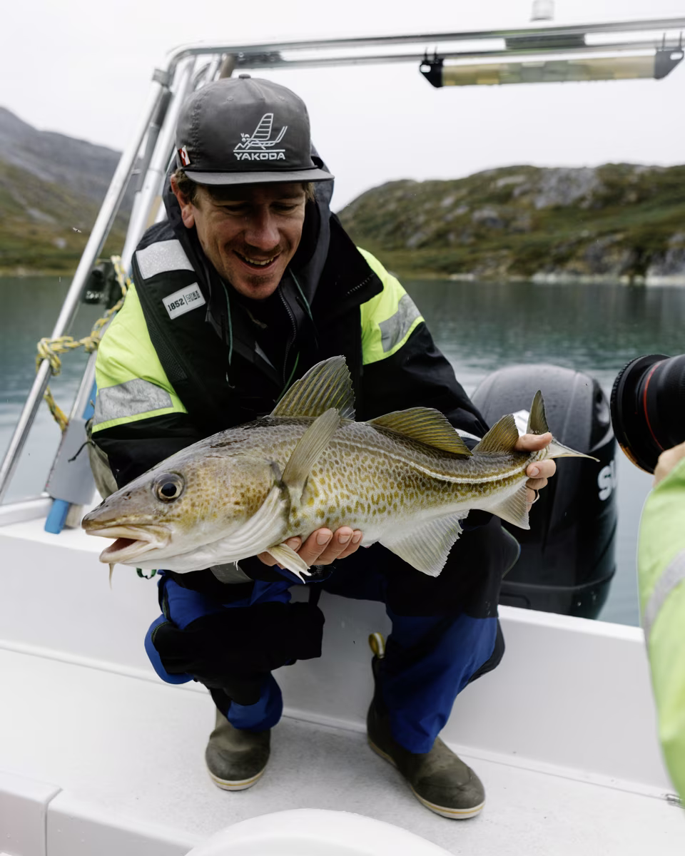 Man holding a large fish on a boat with water and hills in the background.