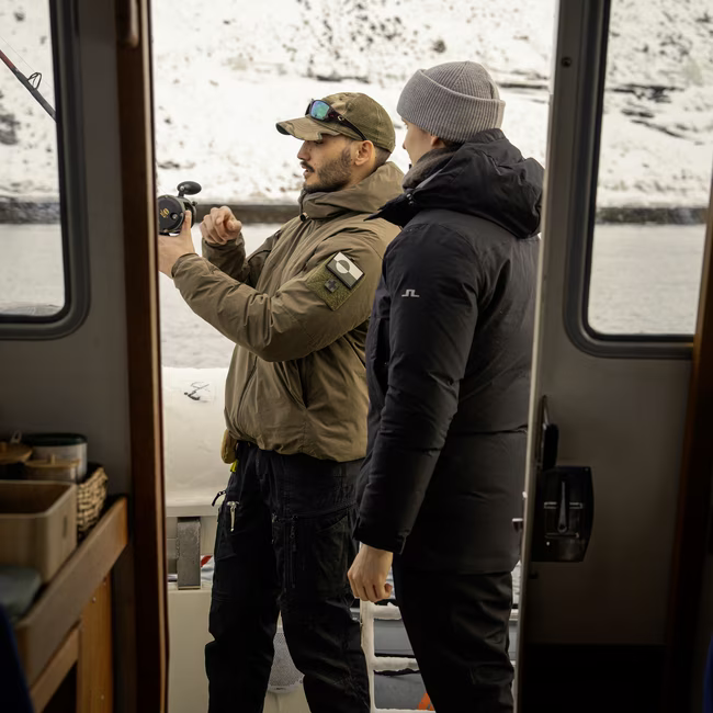 Two men on a boat preparing a fishing rod with snowy landscape in the background.