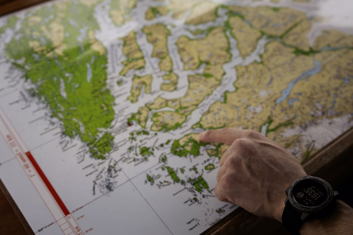Hand wearing watch pointing at a green and yellow map on a wooden table.