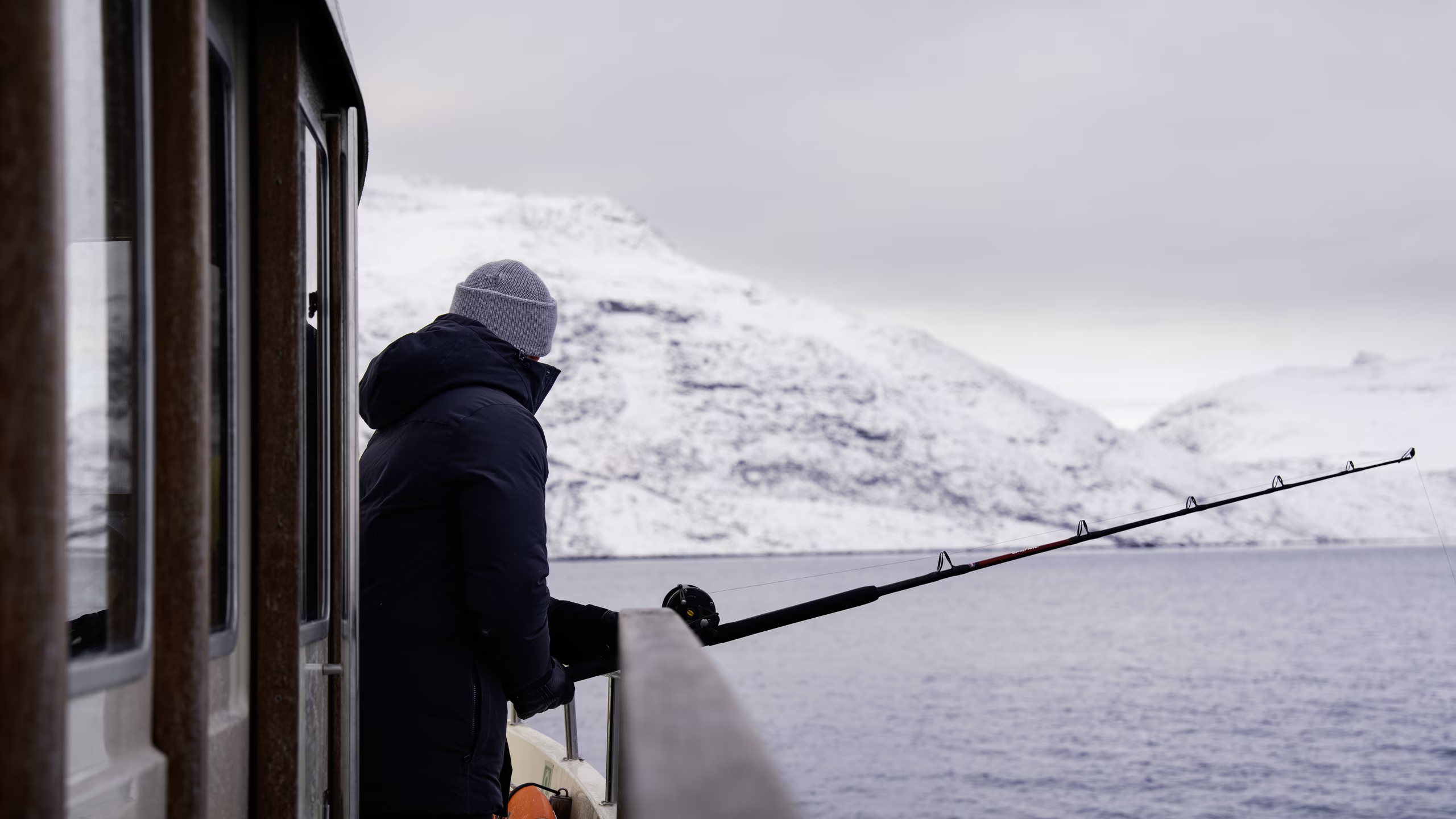 Person wearing a beanie and jacket fishing from a boat with snowy mountains in the background.