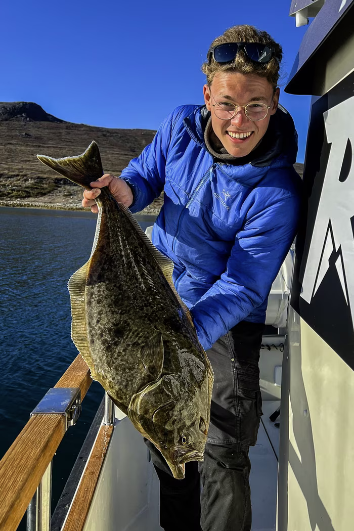 Smiling person in glasses and blue jacket holding a large flat fish on a boat with water and hills behind.