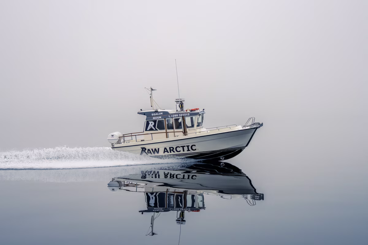 Boat named RAW ARCTIC speeding on still water with clear reflection under a gray sky.