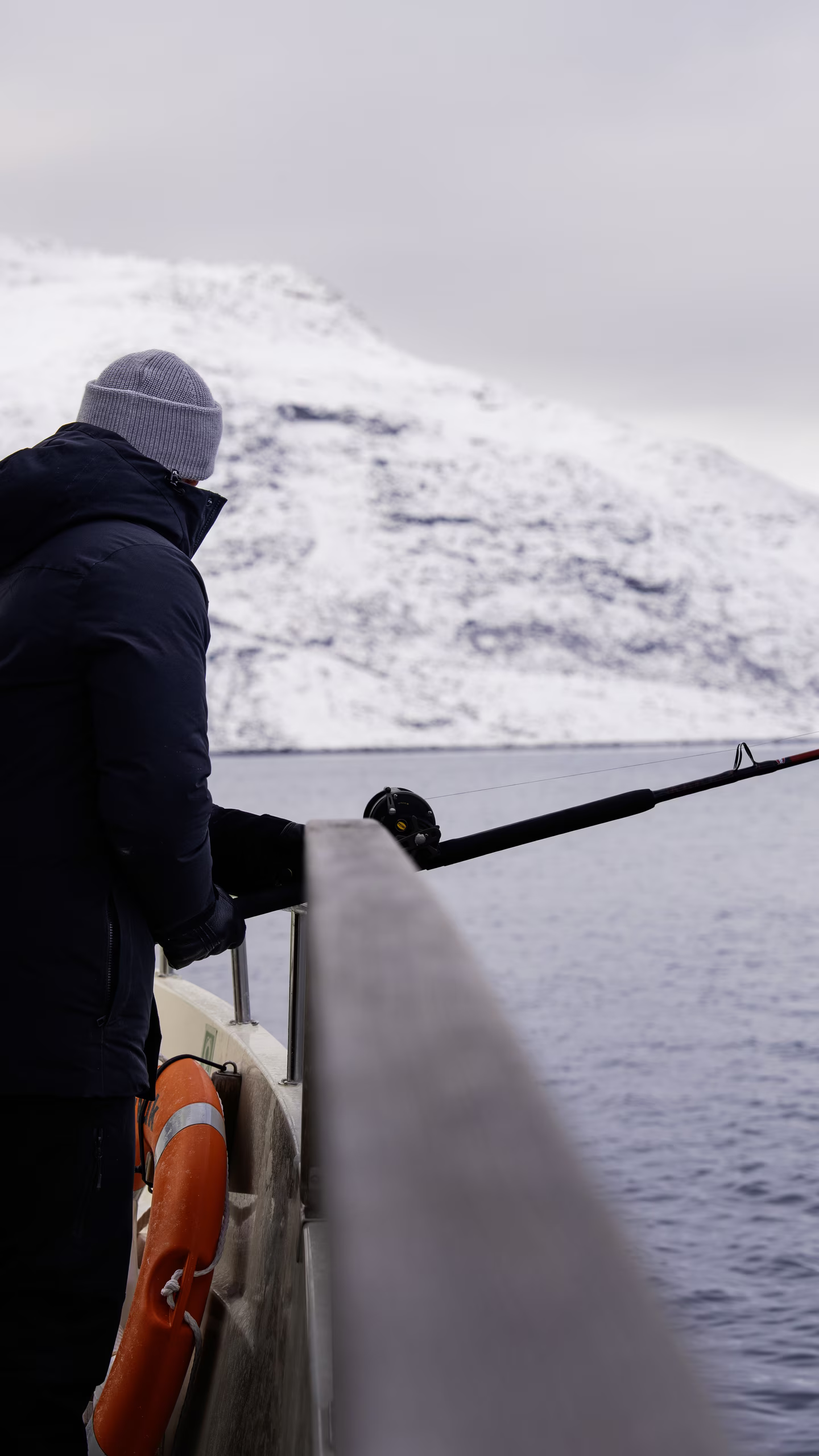Person fishing from boat wearing winter clothing with snowy mountains and water in background.
