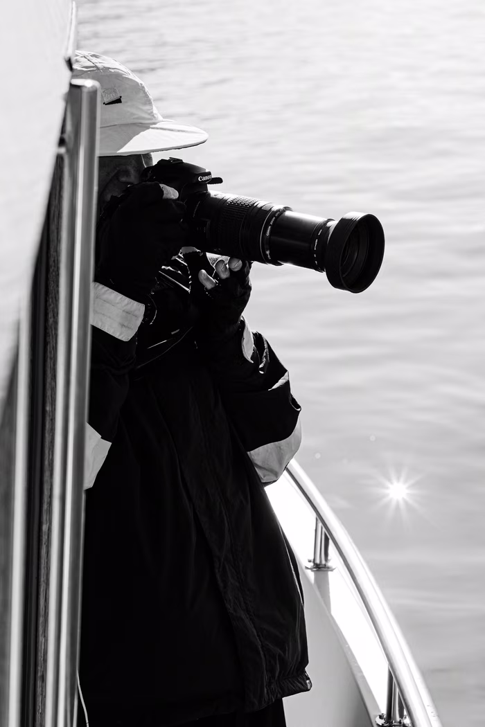 Person in a hat and jacket taking photos with a telephoto lens camera from a boat.