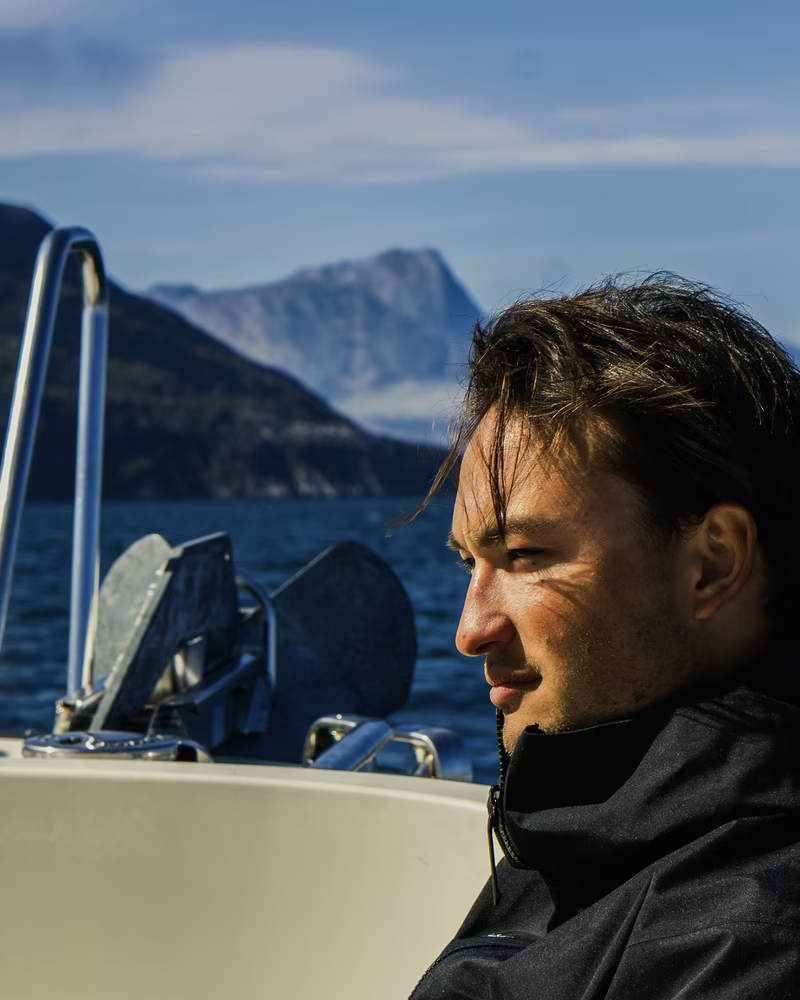 Man in black jacket sitting on a boat with mountains and water in the background.