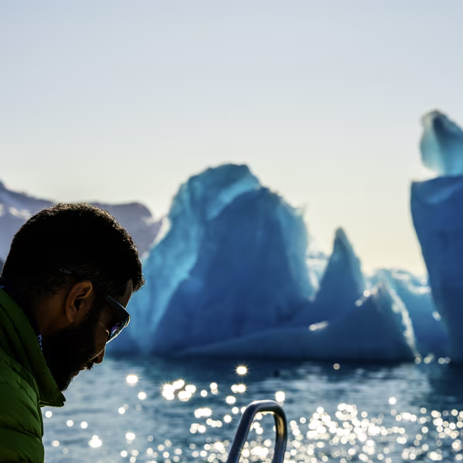 Man wearing sunglasses and a green jacket on a boat with large icebergs in the background.