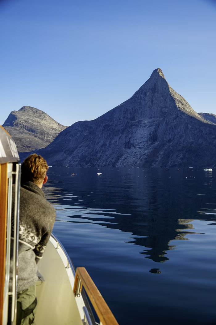Person on boat looking at a sharp mountain peak reflected in calm water under a clear blue sky.