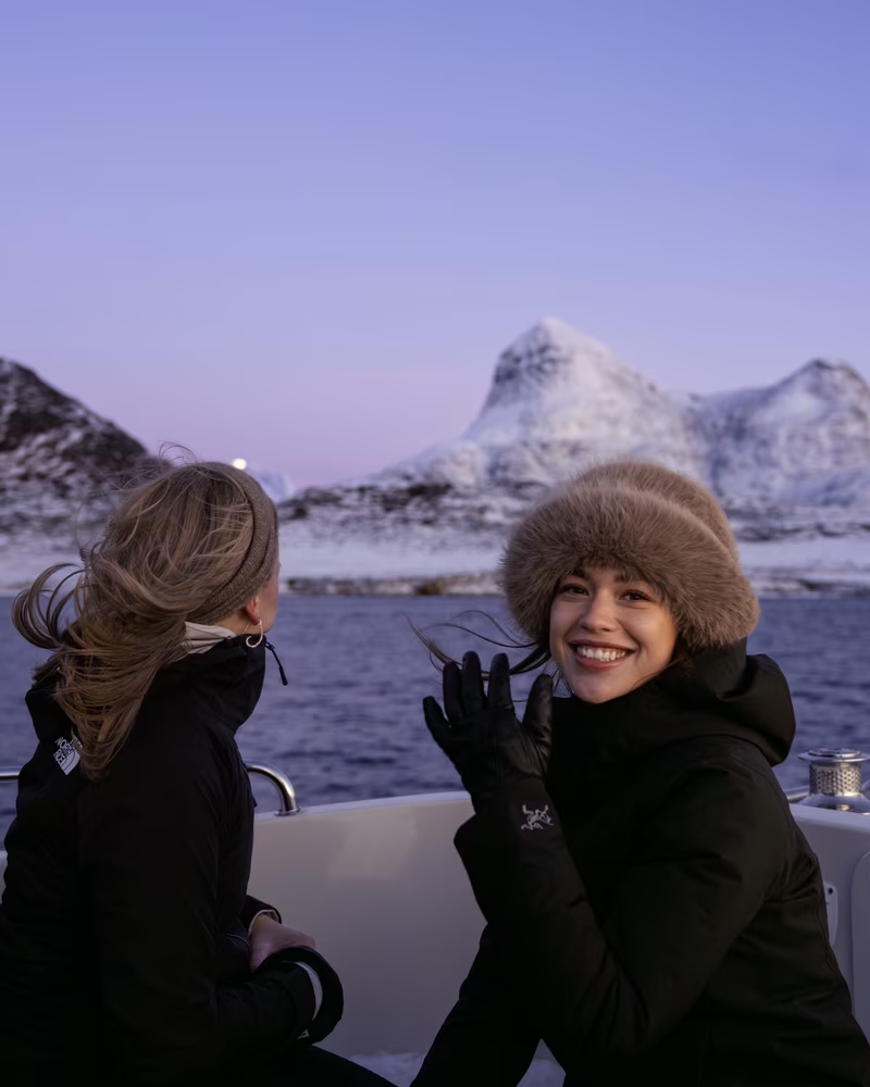 Two women in warm jackets on a boat with snowy mountains and water in the background at dusk.