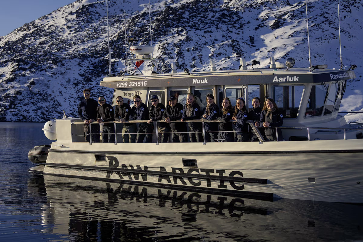 Group of people standing on a boat named Raw Arctic with snowy mountain in the background.