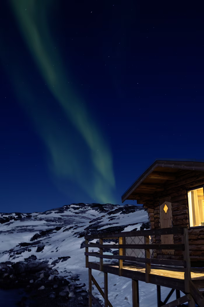 Wooden cabin with lit windows on snowy mountain at night under the northern lights.