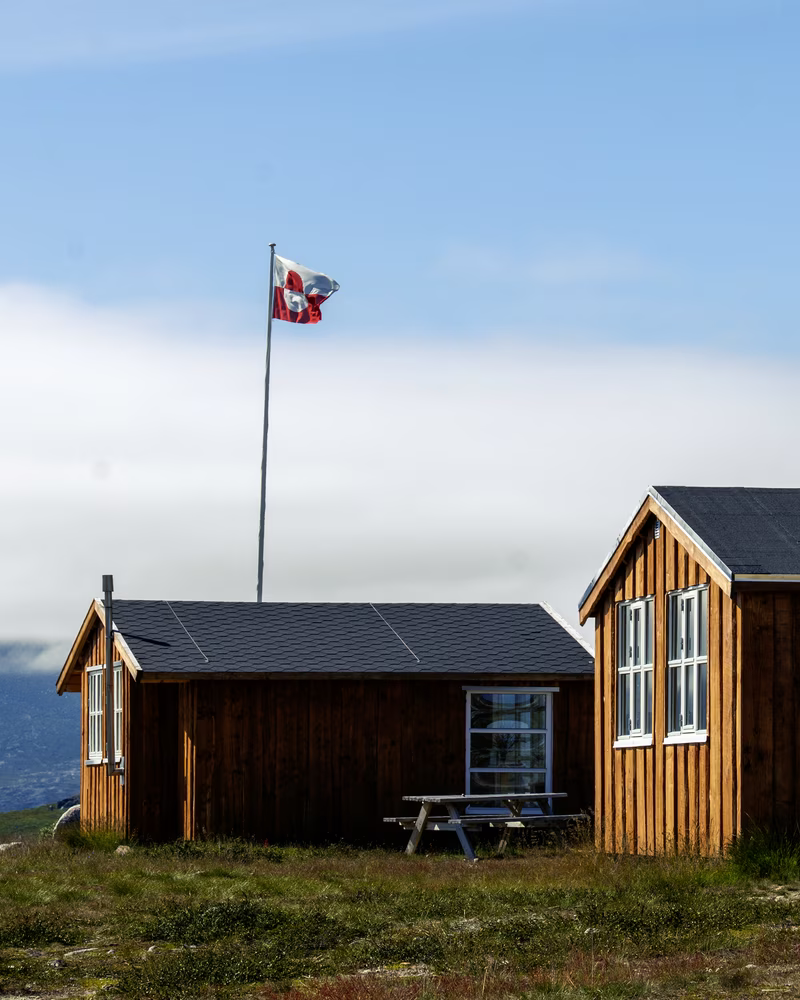 Two wooden cabins with large windows on grass under a blue sky with a flagpole flying a red and white flag.