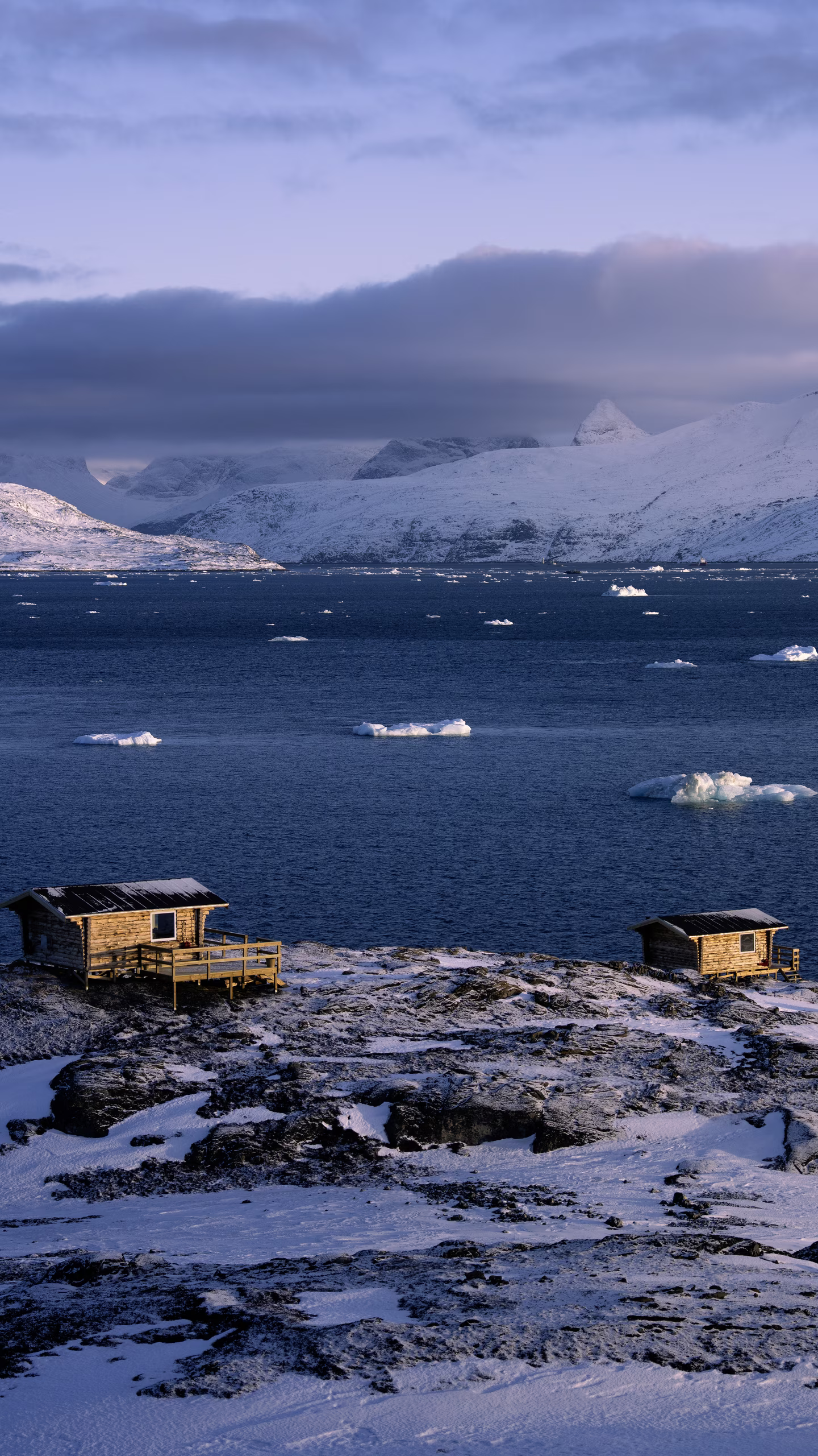 Two wooden cabins on snowy rocky shore with floating ice chunks and snow-covered mountains in the background.