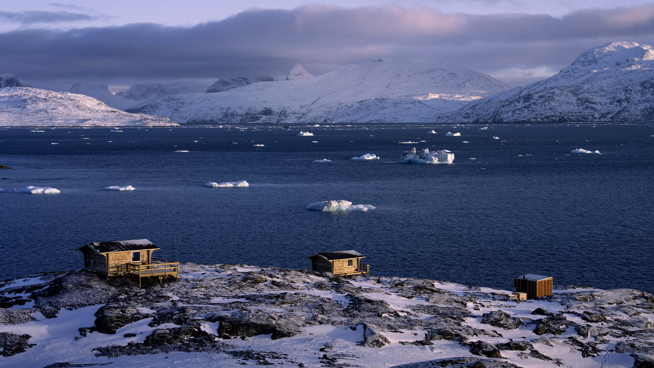Two wooden cabins and a shed on snow-covered rocky shore by icy sea with snowy mountains beyond.