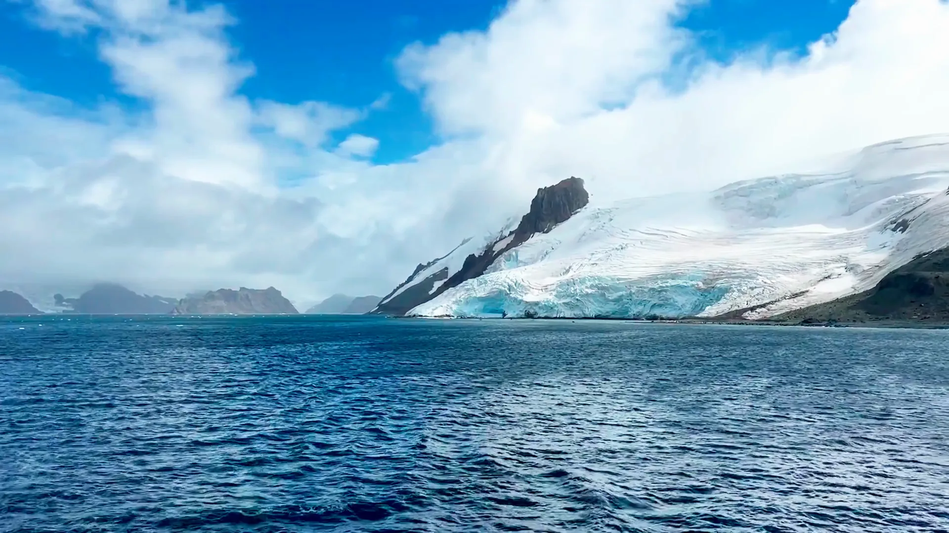 Snow-covered mountain beside a deep blue ocean under a partly cloudy sky.
