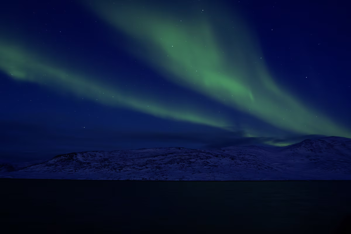 Green northern lights shining over snowy mountainous landscape under a starry night sky.