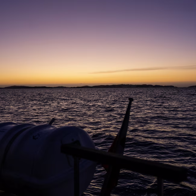 Sunset view over rippling ocean waters with distant islands and boat railing in foreground.