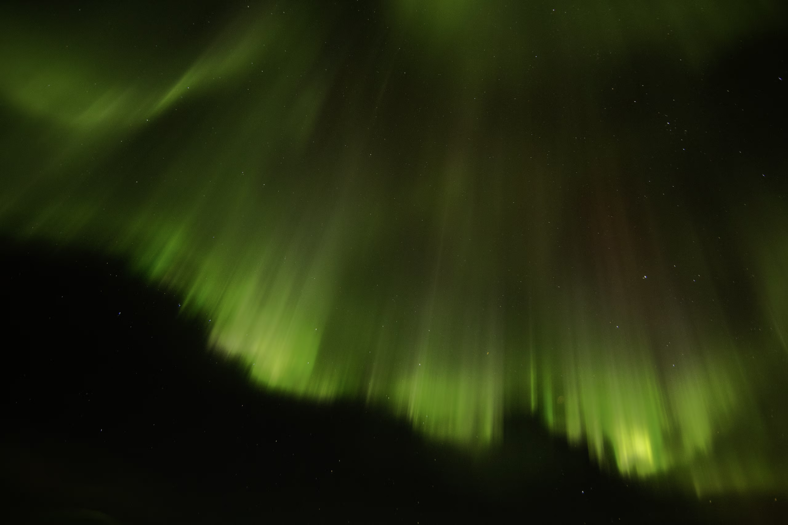 Green northern lights glowing and streaking across a starry night sky above dark silhouette hills.