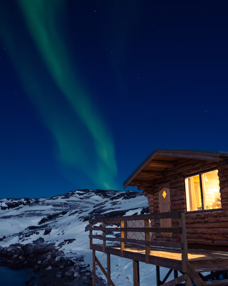 Wooden cabin on stilts with lit window under green northern lights in a snowy mountainous landscape at night.