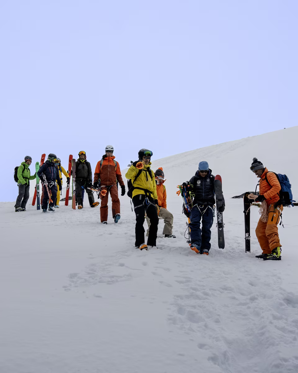 Group of skiers in colorful gear walking on snow carrying skis and poles under clear sky.