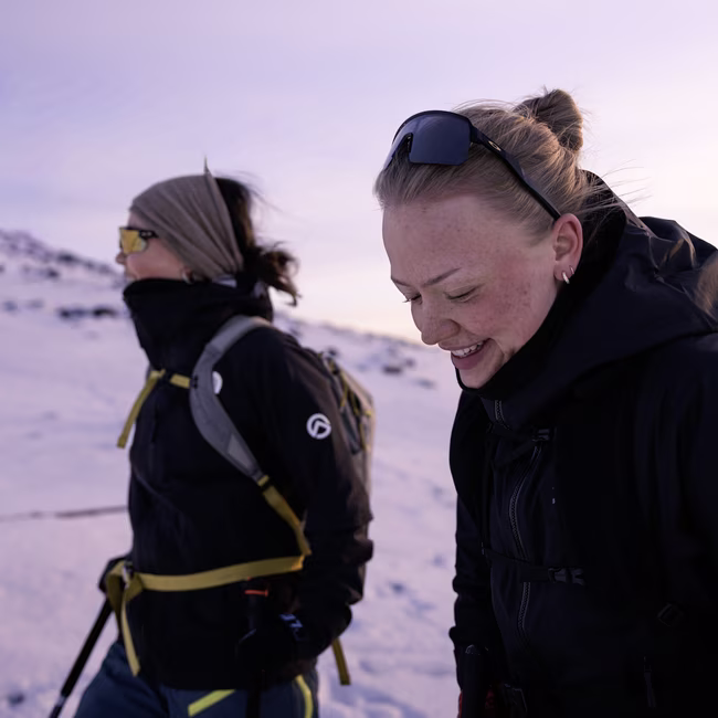 Two women dressed in winter gear outdoors, one smiling and looking down, snowy landscape background.