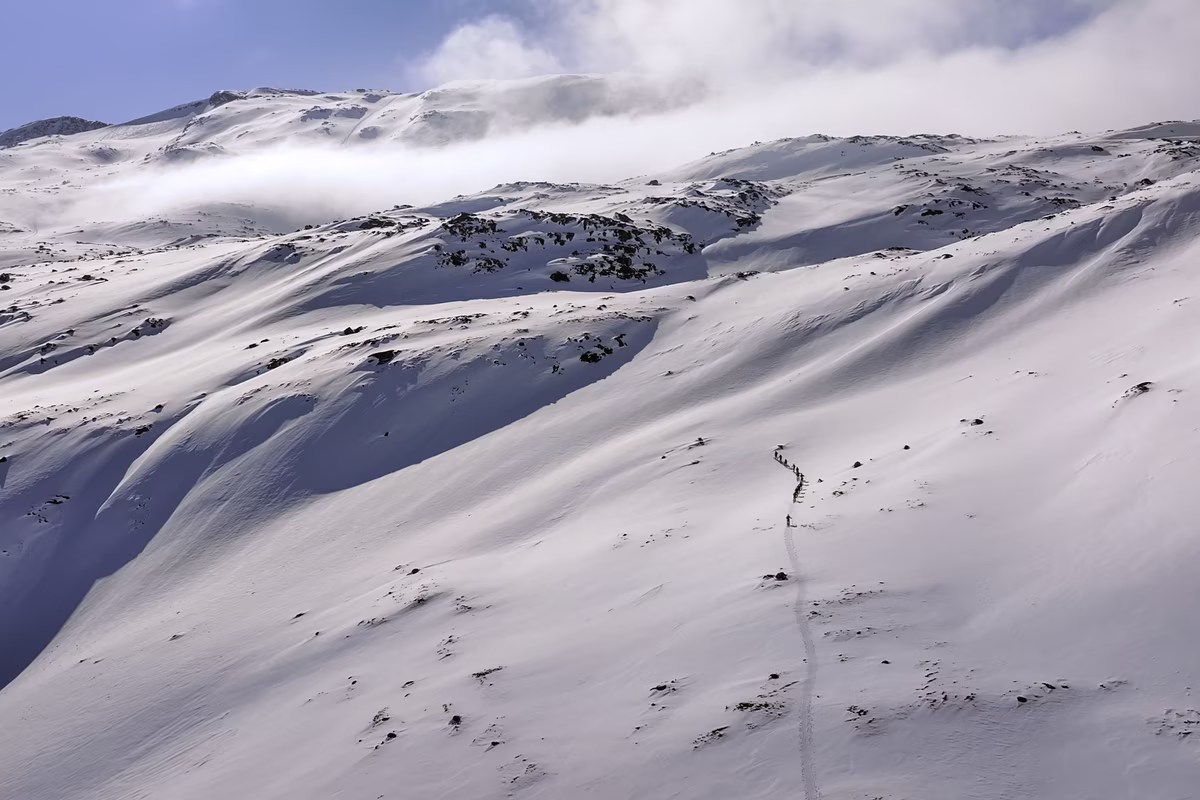 Remote group hiking in single file on a snowy mountain slope under a partly cloudy sky.
