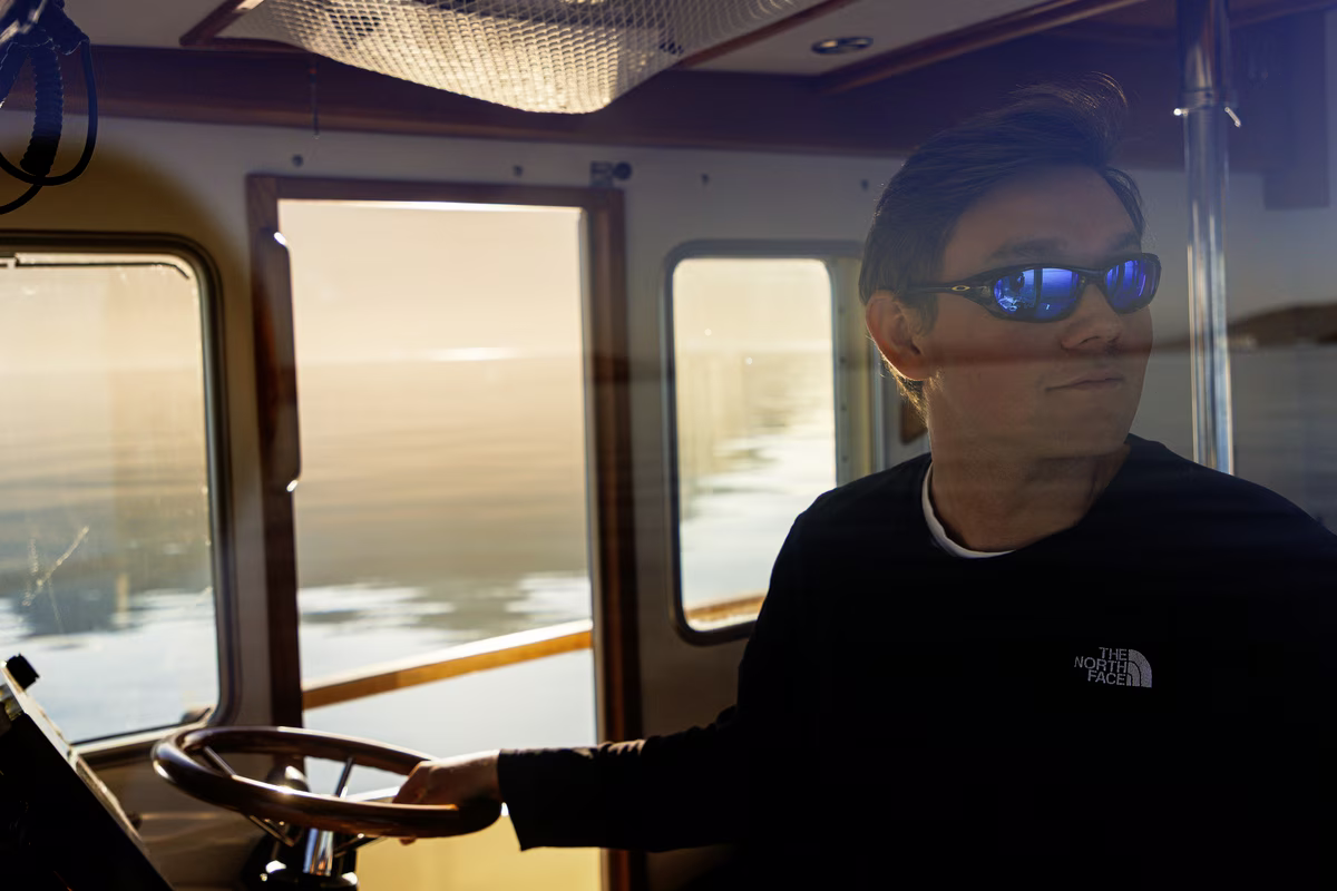 Man wearing sunglasses steering a boat inside the cabin with water visible outside windows.