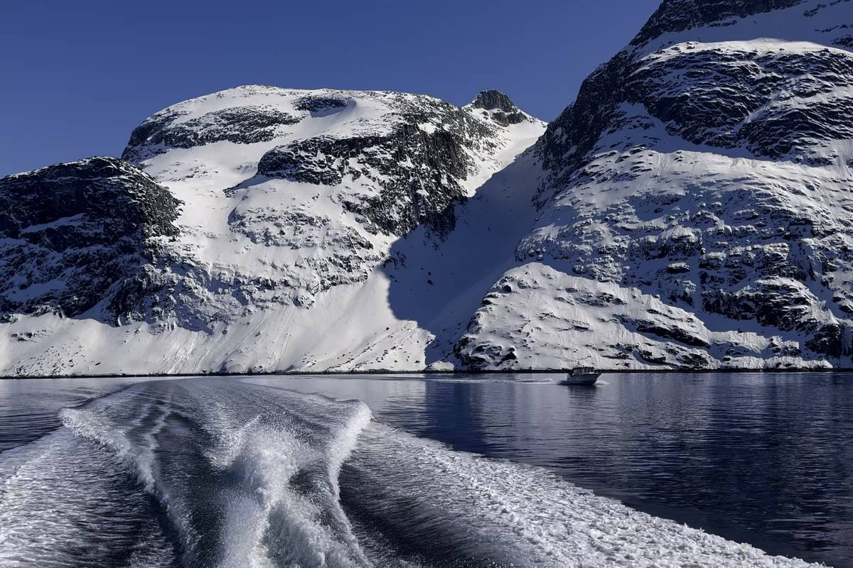 Wake of a boat on calm water with snow-covered rocky mountains under a clear blue sky.