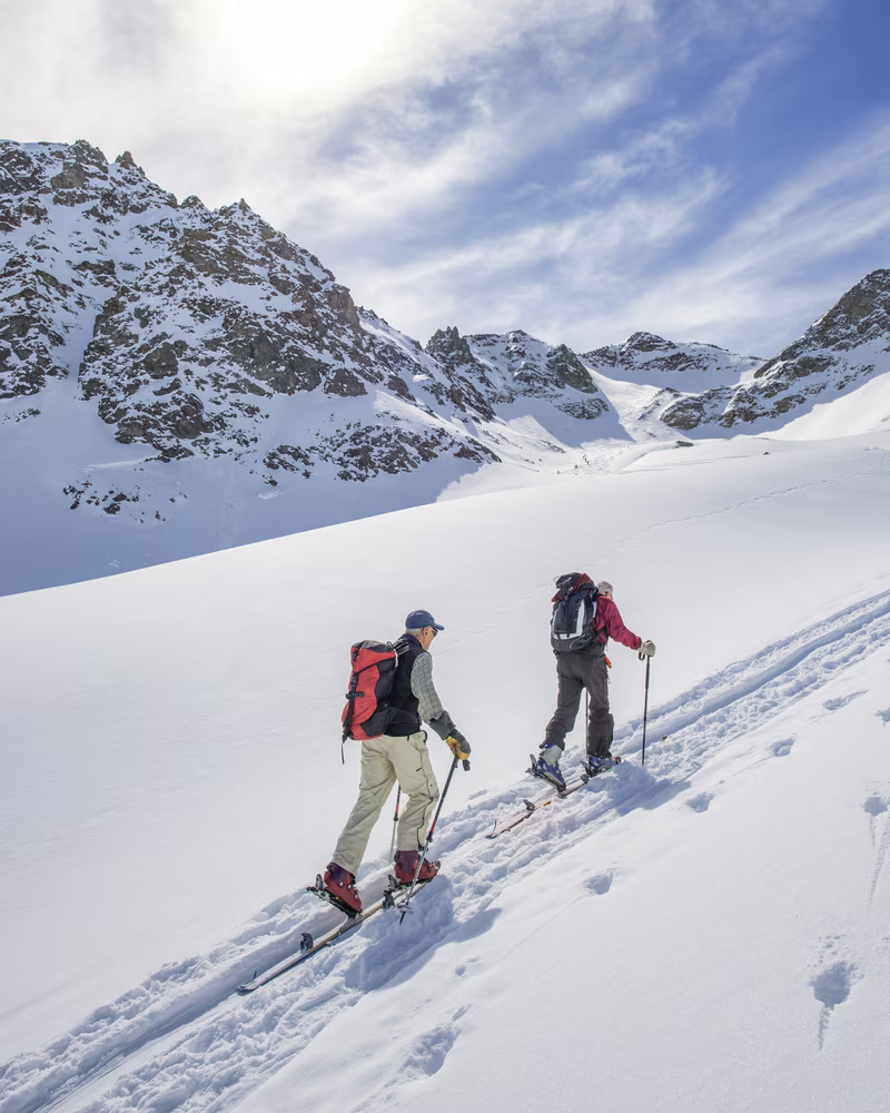 Two people skiing uphill on a snowy mountain slope with rocky peaks in the background.