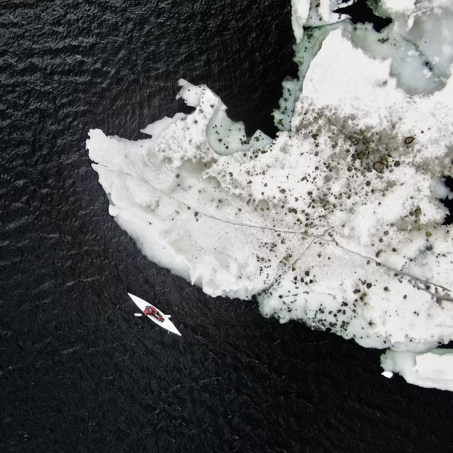 A person in a white kayak paddling near a large ice formation in dark water from above.