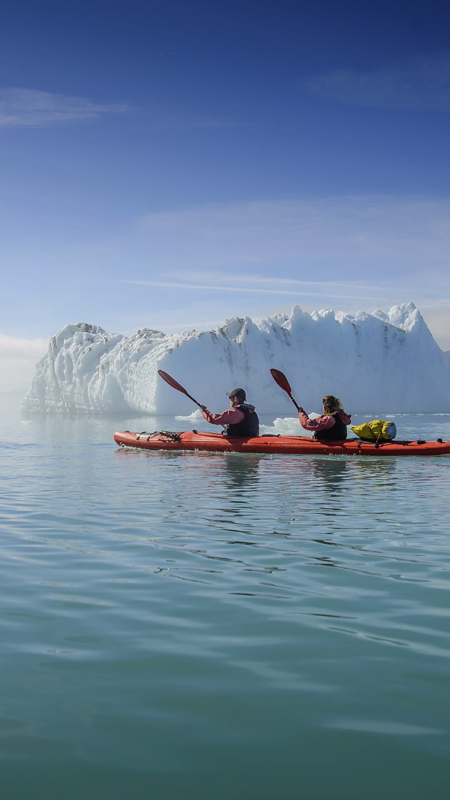 Two people kayaking on calm waters near a large iceberg under a clear blue sky.