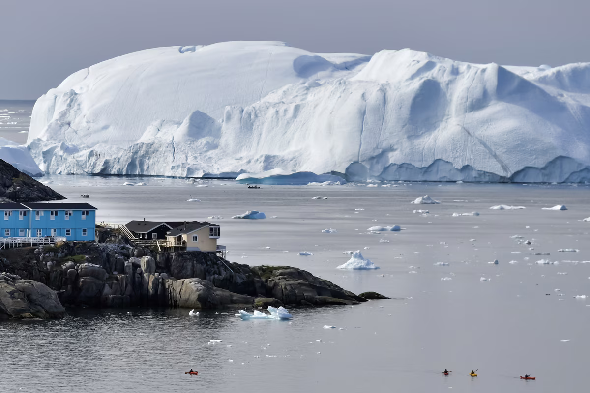 Four kayakers paddle near rocky shore with houses, large iceberg, and scattered ice in calm water.